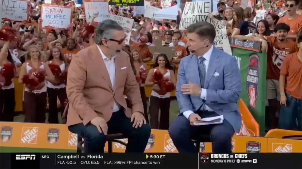 A photo of Senator Ted Cruz (left) is wearing an orange suit (the color of the Texas Longhorns). He has a television ear and mic in his right ear and is speaking to Rece Davis (right). Dozens of Texas Longhorns fans are shown in the background.