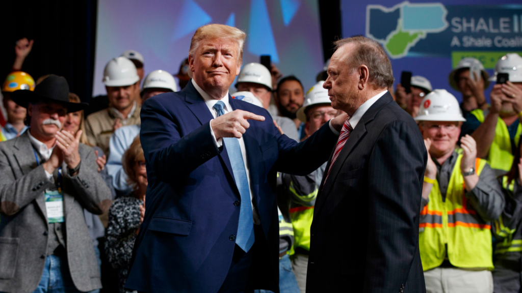President Trump points with both hands at Harold Hamm. He is looking forward and smirking. Hamm is standing to Trump's left and is facing Trump. Over a dozen people, most of whom are wearing worker vests, are standing behind the pair and applauding.