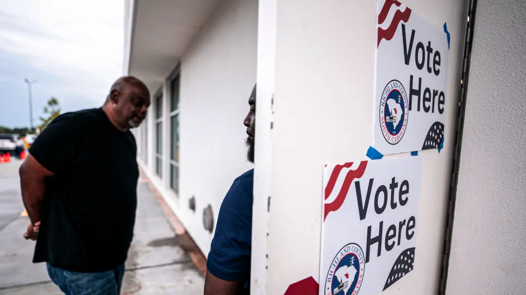 A photograph of a Black man with a gray goatee, wearing a black T-shirt and jeans walking into a polling station. Another Black man stands with his back against the door as he holds it open for the older man. On the other side of the door are two signs that read 