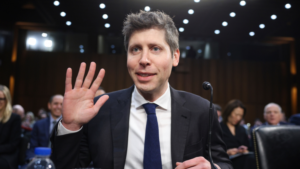 Sam Altman is sitting at a Senate committee hearing wearing a black suit. A microphone is in front of him and he is raising his right hand at shoulder height. Several people are seen sitting in the background.