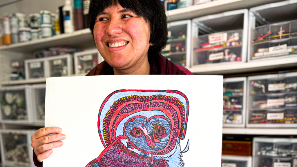 A middle-aged woman with short dark hair smiles as she holds an intricate, colorful drawing of an owl on white paper in front of her. The owl is colored in shades of blue, red, purple, and pink.