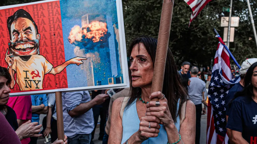 A photo of a middle-aged woman with intense, watery eyes holding a sign. Beside her, someone else holds a sign that illustrates the Mayor of New York laughing while pointing at an explosion at one of the World Trade towers. Others in the crowd also hold signs and American flags.