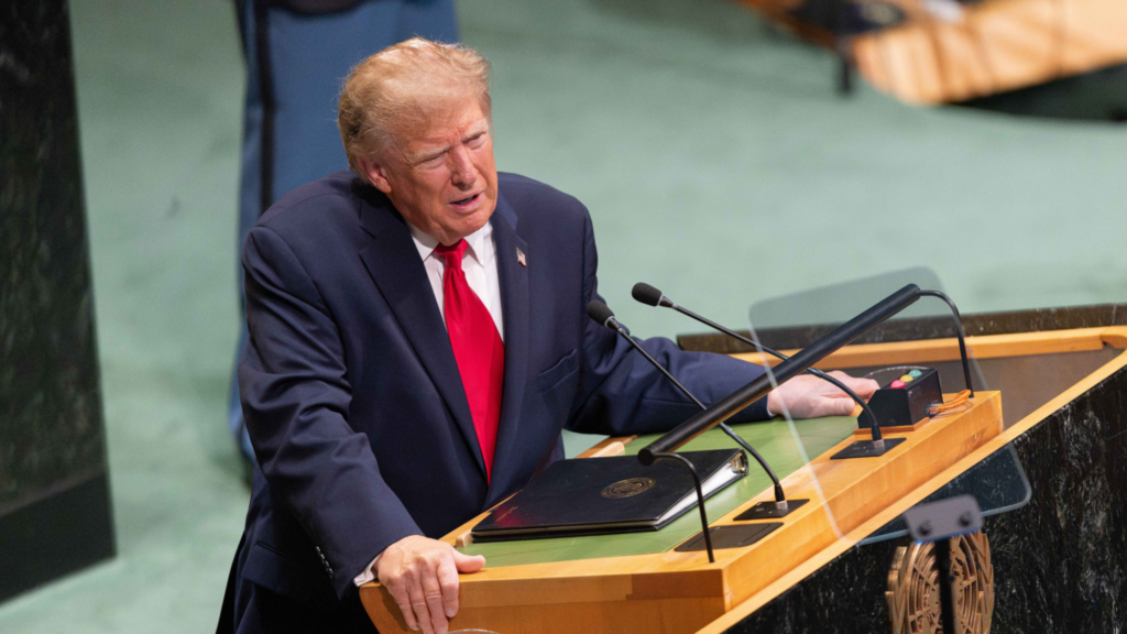 Donald Trump is speaking at podium into a microphone. His head is tilted slightly to the right as he speaks. A teleprompter is shown in front of the podium. He is indoors at the UN headquarters.