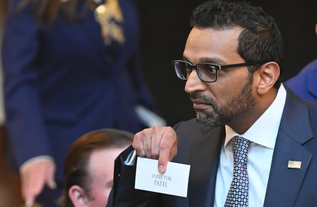 Kash Patel is wearing black glasses and is holding a white card with the words