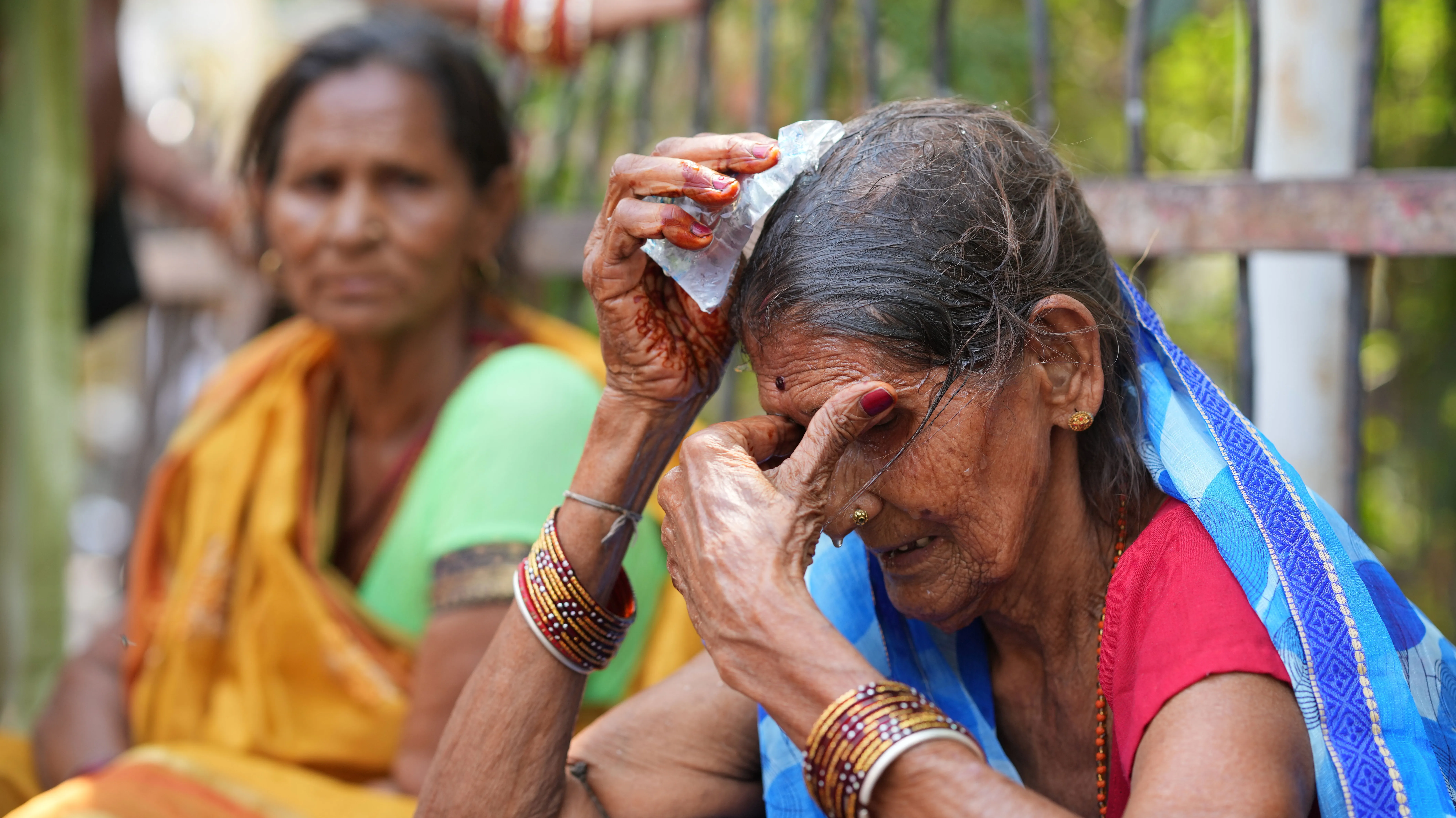 A somewhat frail-looking older woman in a red shirt and blue sari, bangles on her arms, hold an ice pack to her damp hair as another woman in an orange sorry looks on with concern.