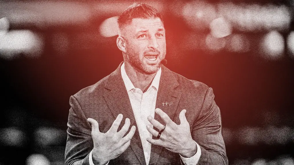 Tim Tebow wearing a suit and speaking into a headset at a football game.