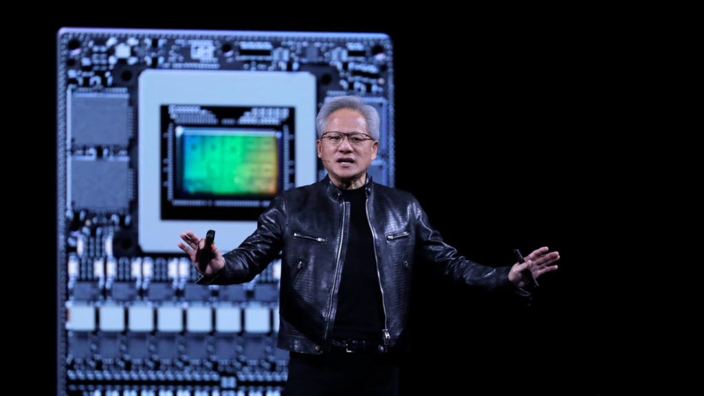 Nvidia CEO Jensen Huang is giving a speech. He is wearing a black leather jacket and black pants. He has an earset microphone in his right ear. In the background is a graphic of what appears to be a GPU chip.