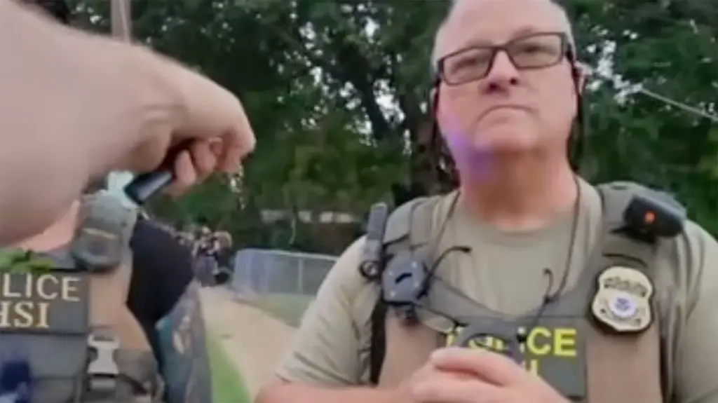 An ICE officer with white hair, probably in his sixties, clad in the usual gear vest and glasses stares at the person filming him and pointing a finger far behind him to where an arrest of an immigrant is taking place.