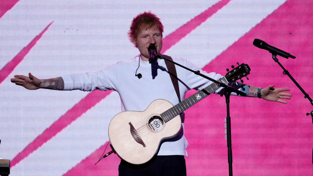 Ed Sheeran is standing with his arms out beside him at shoulder length. He is performing with a microphone in front of his mouth and a guitar strapped to his shoulder.
