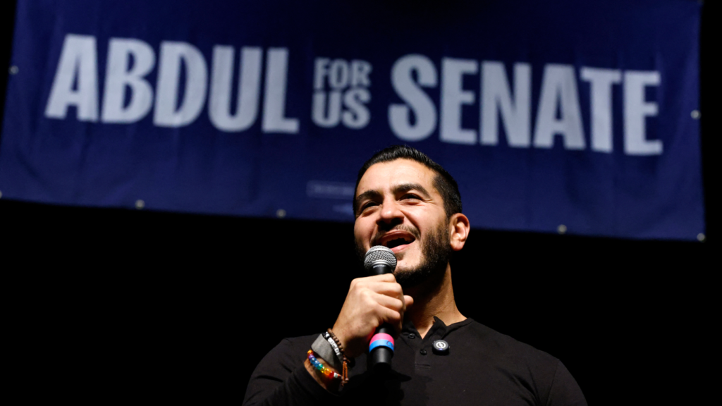 A photo of Abdul El-Sayed holding and speaking into a microphone in his right hand. He is wearing multiple bracelets on his right wrist and is wearing a black shirt. Above him is a sign that reads 