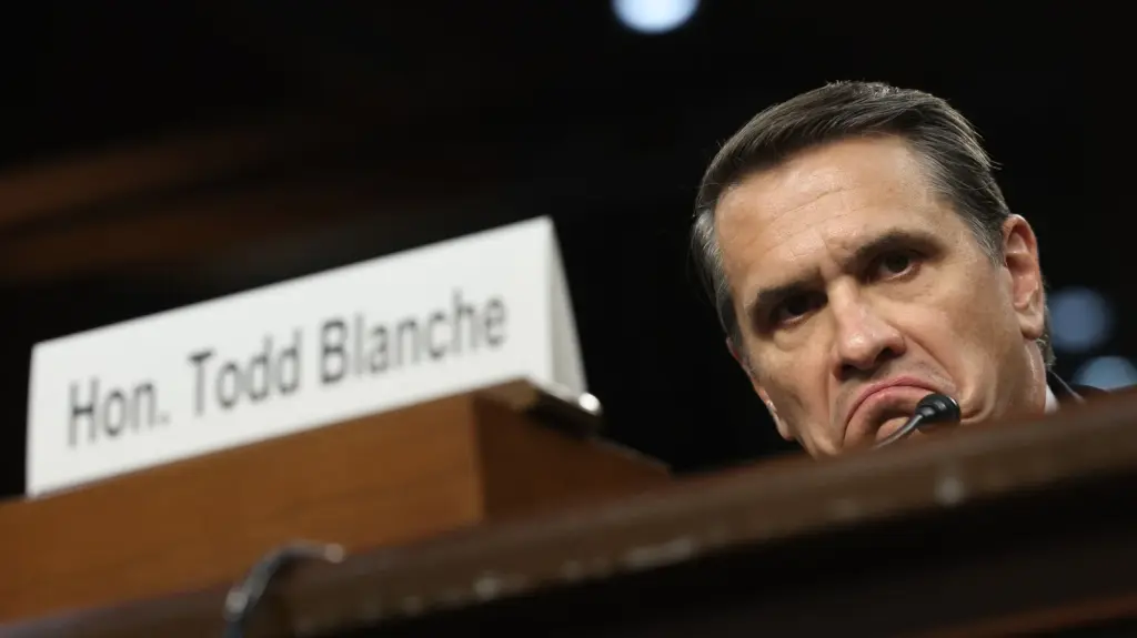 Todd Blanche's face, weathered and gray, peeking over a podium behind a card that says 