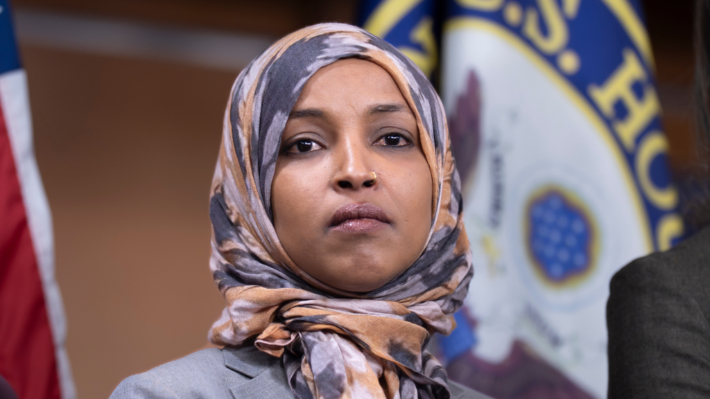A photo of Ilhan Omar that shows her face and shoulders. She is wearing a gray suit and a multi-colored hijab (brown, light gray, and dark gray). Behind her is the flag for the US House of Representatives.