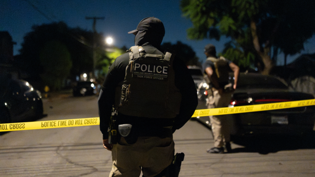 A photo of the back of an officer wearing a black baseball cap, black mask, and tactical vest standing behind yellow police tape outside on a street. There is another officer ahead of him and the tape wearing similar clothes. They are both looking to the left.