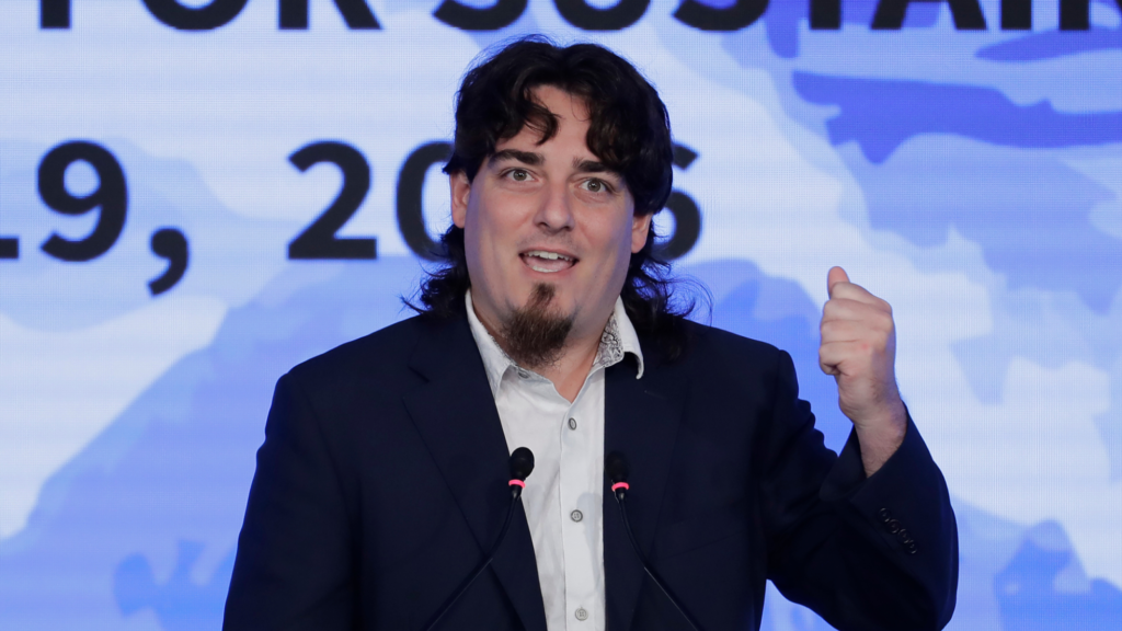 Palmer Luckey speaks into a microphone. He has shoulder-length dark brown hair and a goatee. He is raising his left hand in a fist at shoulder length.