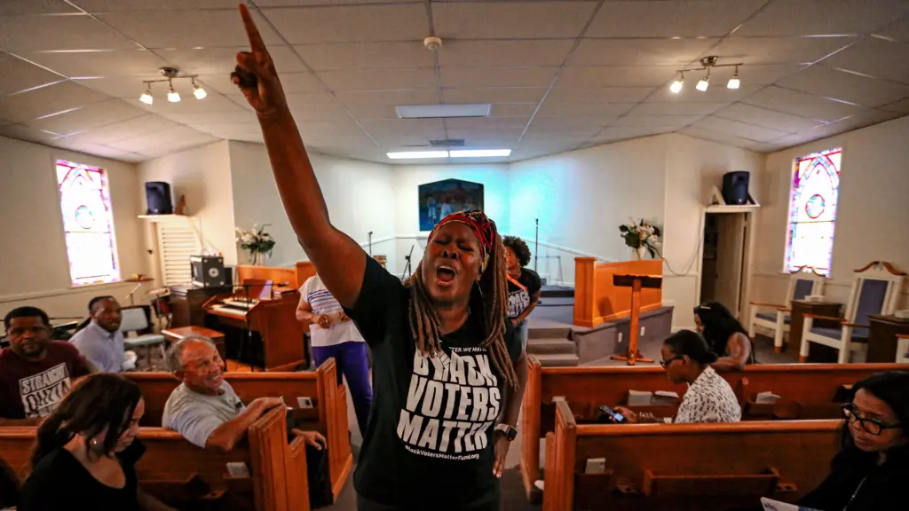 A young Black woman stands in the aisle of a church with her right hand in the air as she speaks with animation. Casually dressed people sitting in the pews to her left and right turn in their seats as they look at her. The woman is wearing a black T-shirt with the words “BLACK VOTES MATTER” in white sans serif type.