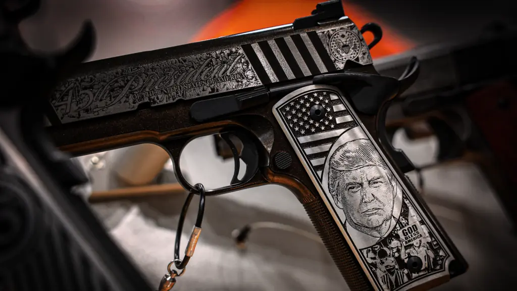 A pistol that has and engraved likeness of Donald Trump's face on the handle with an American flag above and a crowd with a person holding a sign that reads 