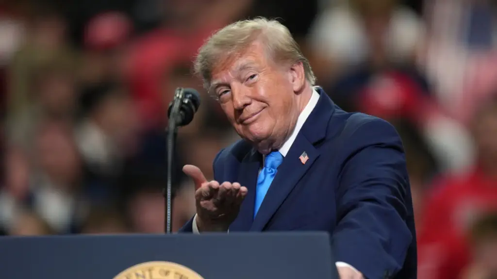 President Trump, in a dark blue suit, blue tie, and a white shirt, with an American flag pin on his right lapel. He's smirking and look directly into the camera, with his left hand pointing toward the camera with his palm facing up. He's standing behind a podium with a mic in the frame.