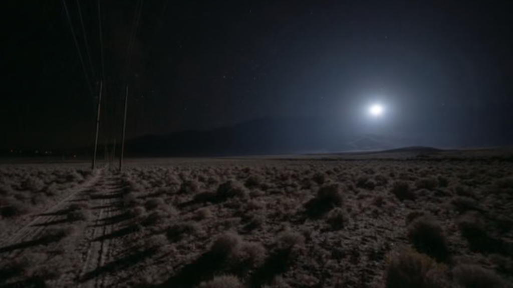 A dark landscape with some shrubs and hills and electric lines, but, most prominently, a very bright object that looks somewhat like the sun in the lower part of the sky on the right.