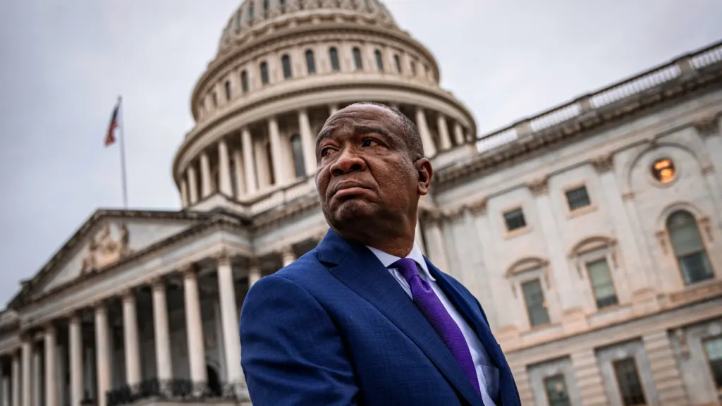A senior Black man in a blue suit and purple tie looks over his right shoulder as the US Capitol building looms in the background.