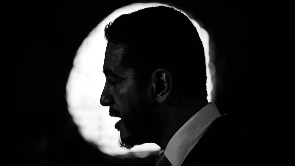 A dark portrait in black and white of a man with a mustache and facial hair on his chin, as seen in profile against a circular bright light. You can make out that he's speaking and wearing a suit and tie.
