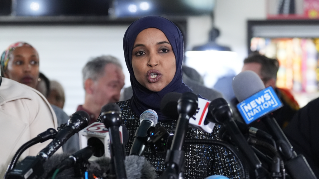 Photo of Ilhan Omar speaking at a press conference. Several microphones displaying logos of news outlets are pointed toward her. She is wearing a black hijab and a black top with white dots scattered across it. Several people stand in the background.