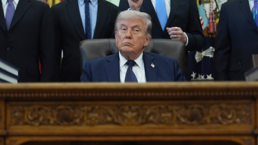 President Trump is sitting in a black chair at a wooden table. Several people are seen standing behind them in the background. We only see their torsos in the photo.