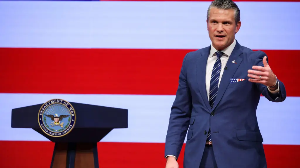 A middle-aged Caucasian stands beside a podium on a stage. He wears a blue suit and Amercian flag-patterned handkerchief in his blazer's breast pocket while gesturing with his left hand.