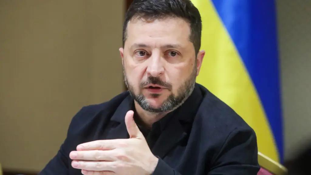 A white man with brown hair and short beard sits in front of a Ukrainian flag and gestures with a hand.