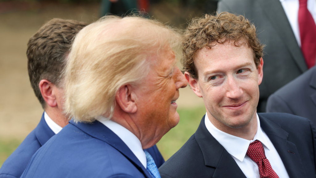 Mark Zuckerberg looking at Donald Trump and smiling. Trump is wearing a blue suit and Zuckerberg is wearing a black suit. We see both men from the shoulders up. Three other people are seen in the background but are cut off by the photo.