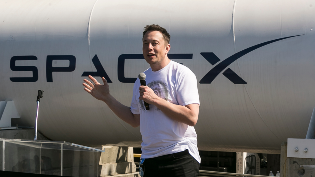 Photo of Elon Musk speaking into a microphone that he is holding in his left hand. He is wearing a white shirt with a gray graphic design on the upper half of the front. Behind him is a large white structure with the word SPACEX in black.