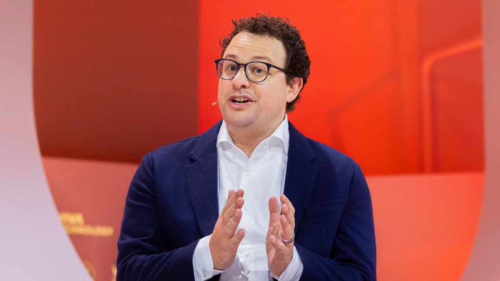 A photo of Dario Amodei wearing a dark blue suit, white colored shirt, and black glasses. We seem him from his chest up and he is speaking into an earpiece microphone at a tech conference.