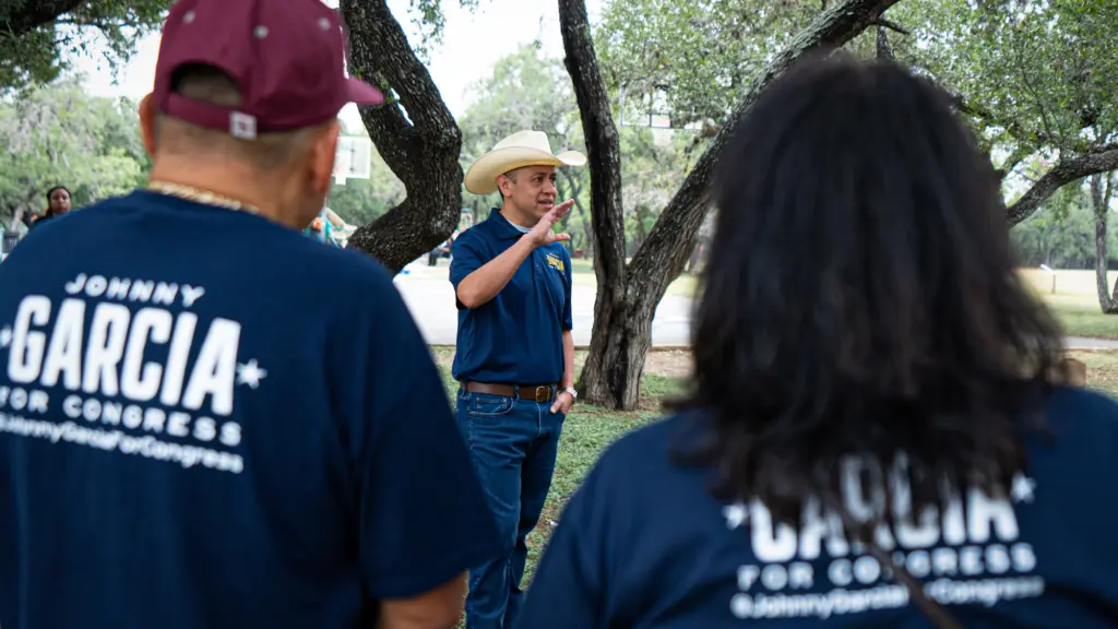 A man dressed in a blue polo shirt, jeans and cowboy hat stands in a grove of trees as he addresses campaign volunteers, who are dressed in blue shirts that read