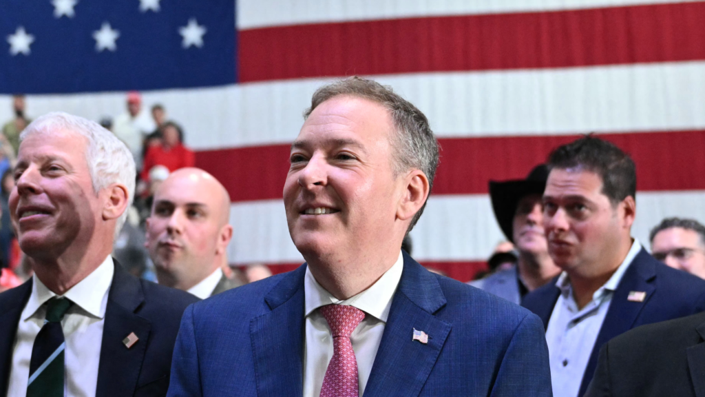 Smiling white man in the center of a crowd of other white men in front of a red, white, and blue flag. They all watch something in the distance.