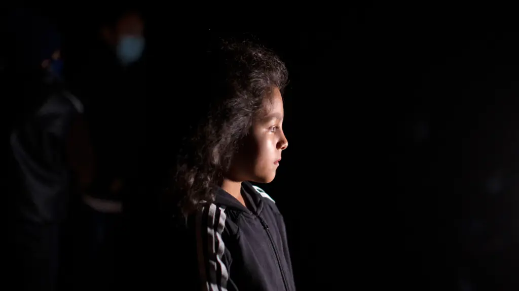 A young girl looks to the side, in front of a dark background.