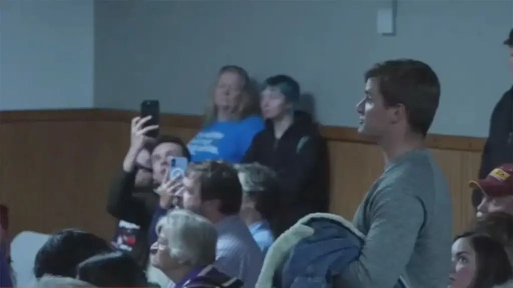 A photo of a left-side profile young white man in a gray long-sleeved shirt facing forward speaking in a town hall event. He is standing while several people in the audience sitting or standing around him.