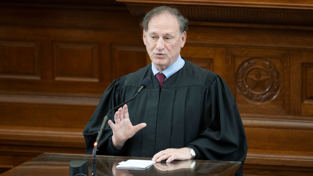 A man in a black justice's robe, blue collared shirt, dark red tie, and wrist watch speaks into a microphone at a podium with notes in front of him.