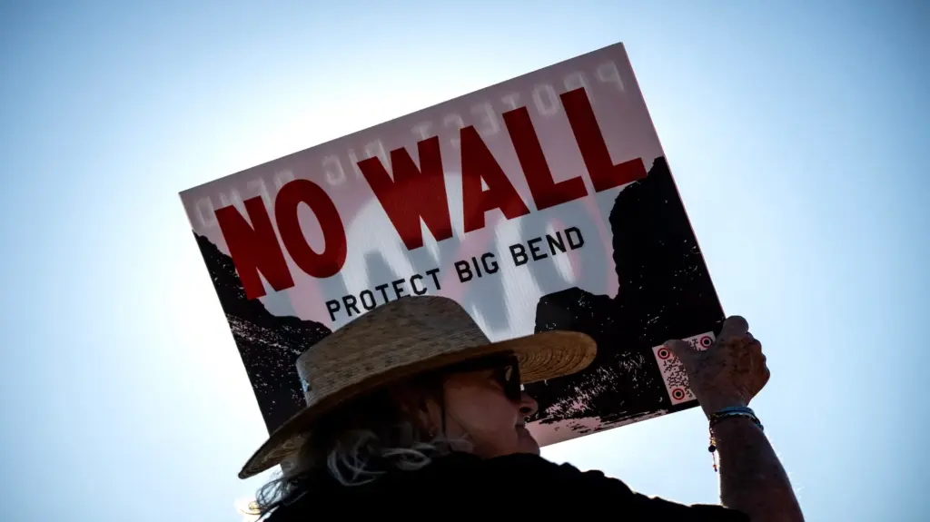 A woman in a wide-brimmed hat holds a sign reading, 