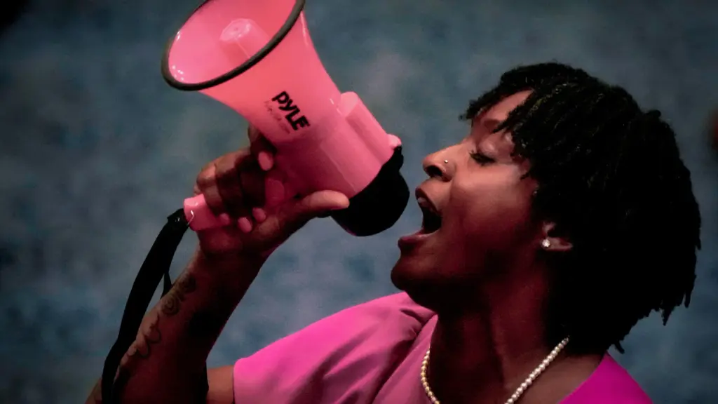 An African American woman with medium-length twist locs shouts into a small pink bullhorn. She wears a pink dress with pearls around her neck.