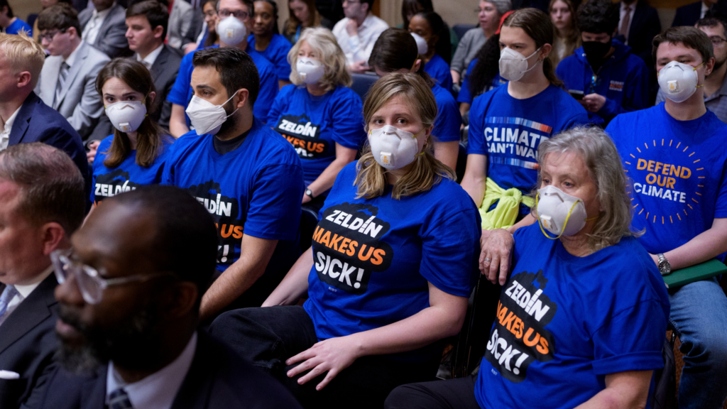 People in white masks and blue shirts sit in rows on chairs in a meeting. Their shirts say