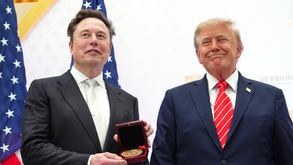 Photo of Donald Trump (right) smiling and standing next to Elon Musk (left). Musk is holding a gold medal in a wooden case in front of himself with both hands. They are both wearing suits. Two American flags and a white wall are seen behind them