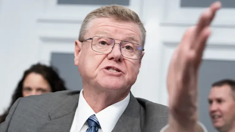 A close up shot of a white man in a gray suit and blue tie and glasses gesturing with his hand as he speaks.