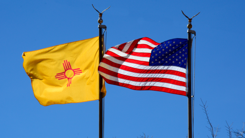 Photo looking up at the American flag (right) and New Mexico state flag (left) flying side-by-side outside. The sky is bright blue and the sun (offscreen) is shining. Some tree branches are seen in the bottom right of the photo.