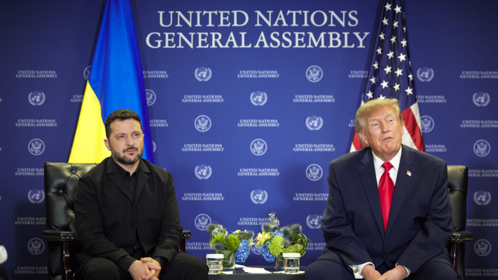 US President Donald Trump and Ukraine President Volodymyr Zelensky are both sitting in black chairs. They are both looking to slightly to their left sides toward something outside of the photo. In the background is one Ukraine, one US flag, and a blue press conference wall that reads 