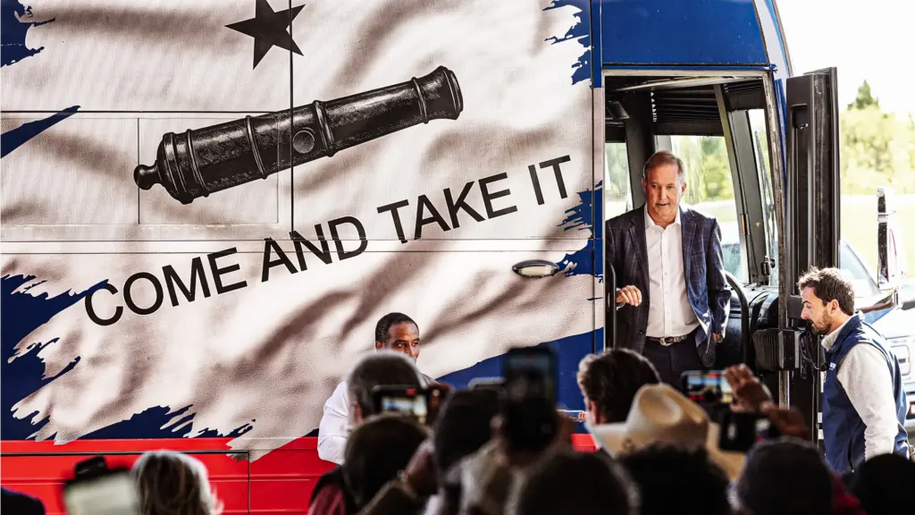 A photo of a middle-aged white man in a suit and white shirt without a tie standing in the door of a boss that has an image of a cannon and star along with the words