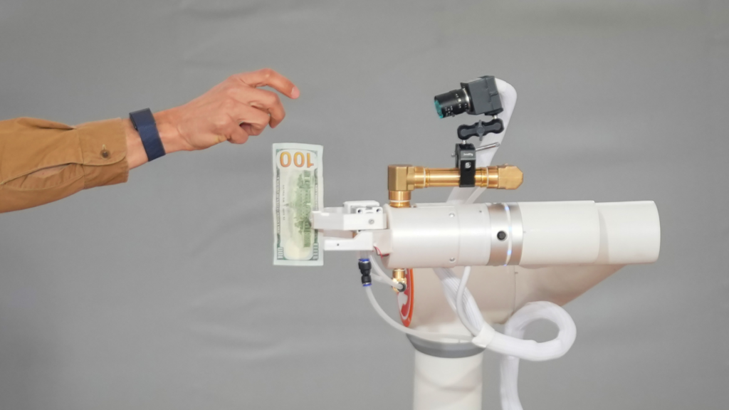 A photo of a person's right arm dropping a $100 bill. We see a brown sieve and a black watch on the person's arm. The robot has a camera-looking device on top and is holding the $100 bill in a white claw-like apparatus.