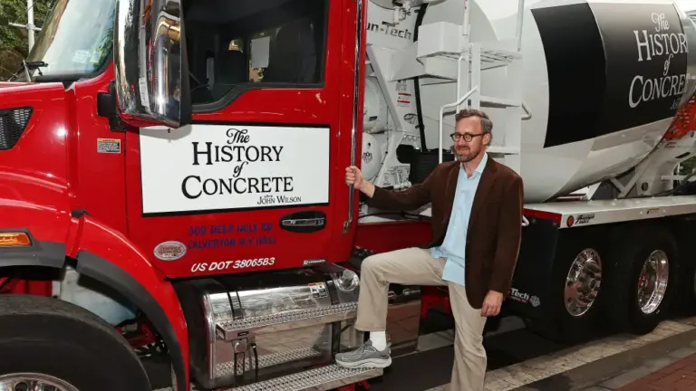a skinny white dude with brown hair, glasses, a beard, in a brown jacket and khaki pants, poses next to a red and grey concrete mixer truck with 'The History of Concrete' label under the driver's side window.