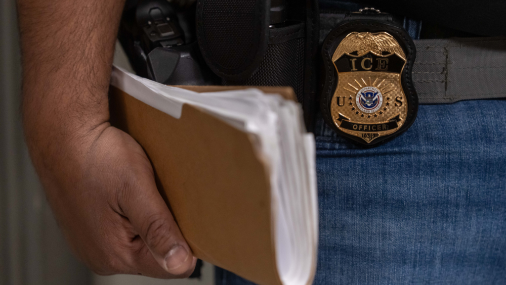 A person is showing holding a brown folder with dozens of pieces of paper inside in their right hand. We also see the person's badge saying